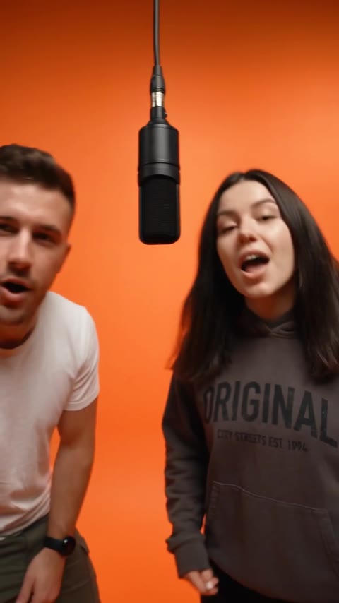 Hotel Lobby AI duo leaning toward the hanging microphone in an orange studio close-up