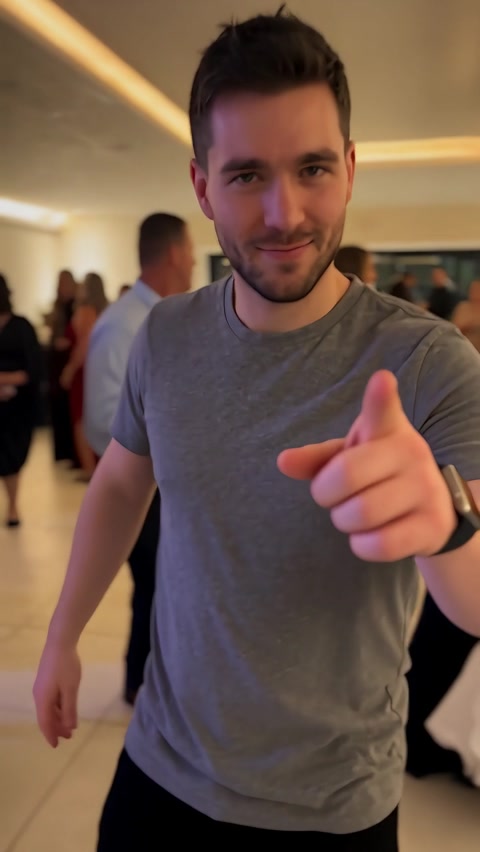 Man in a gray T-shirt pointing toward the camera among banquet guests